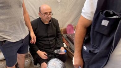 A priest with a bandaged leg sits on a bed inside a tent, surrounded by people offering assistance after an attack on Gaza's Holy Family Church.