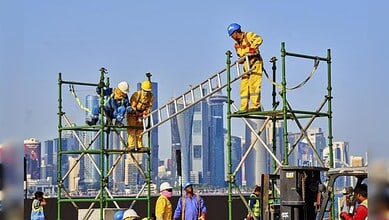 Migrant construction workers wearing safety gear and working on scaffolding with Gulf city skyscrapers in the background.
