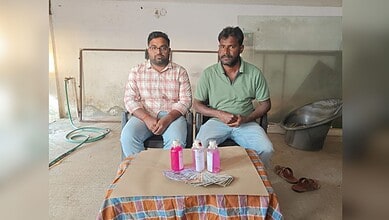 Image shows two people sitting on chairs with cash and bottles with pink liquid in front of them.