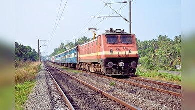 An orange and yellow electric locomotive pulling blue passenger coaches through lush green countryside on an electrified railway track
