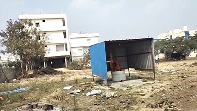 Image shows a vacant urban plot with makeshift shed and residential buildings in background