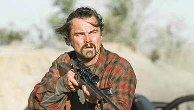 Man with rifle in desert, intense expression, rugged appearance, outdoor setting.