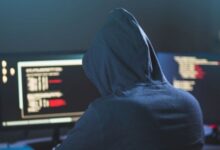 Image shows Person in a hoodie working on a computer with code on multiple screens in a dark setting