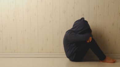 Image shows A person in a dark hoodie sits alone on the floor against a wall, hugging their knees in a posture of distress or despair.