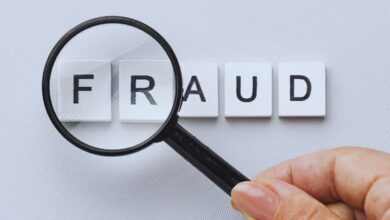 image shows A magnifying glass held over letter tiles spelling "FRAUD" on a light gray background.