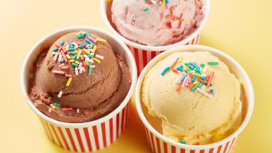 Three scoops of chocolate, strawberry, and vanilla ice cream topped with colorful sprinkles in red-striped paper cups on a yellow background.