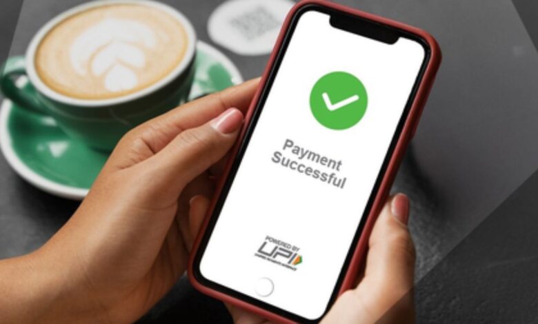 Image shows A hand holds a smartphone displaying a "Payment Successful" confirmation screen powered by UPI, with a coffee cup in the background.
