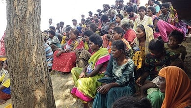 Crowd of men, women, and children sitting on the ground under a tree during a community gathering.