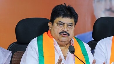 A man speaking at a press conference, wearing a white shirt and a tricolour sash, with a serious expressi.