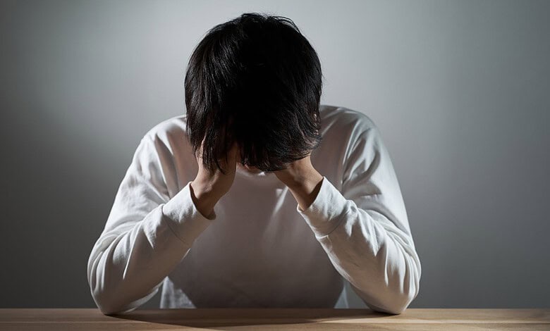 Young man with dark hair sitting at a table, head in hands, experiencing stress or anxiety.