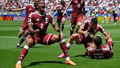 Celebrating a goal, Qatar football team players in maroon jerseys on the field during a match.