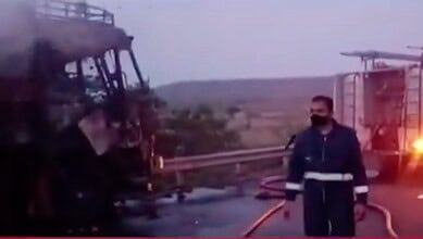 A firefighter inspecting a vehicle fire on a highway at sunset.