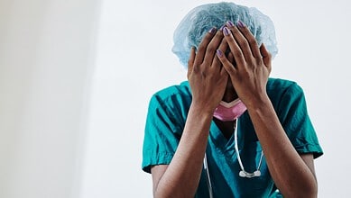 Overwhelmed female doctor with hands covering face in medical scrubs and cap.