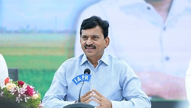 A man in a light blue shirt speaking at a press conference outdoors with a microphone labeled "I & PR".