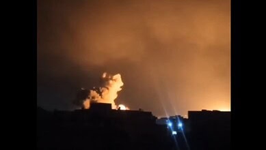 Nighttime scene of explosions and smoke from airstrikes in a cityscape.