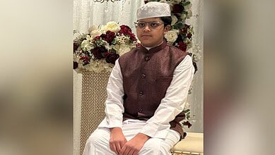 Young boy in traditional Indian attire with glasses and a cap, sitting near a floral arrangement.