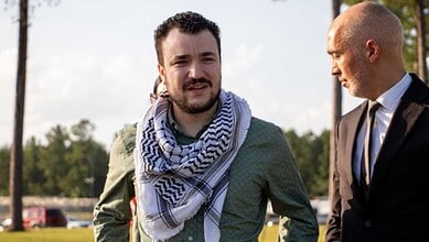 Young man with a beard and scarf talking to a bald man in a suit outdoors.
