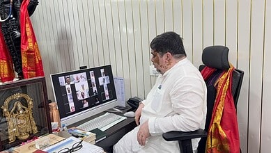 Man in white shirt attending virtual meeting at desk with Gurukul school decor.