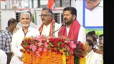 Political leaders at a public event with floral decorations and a large screen in the background.