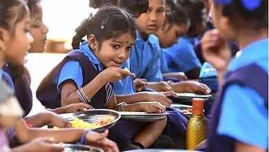 Young schoolgirls in blue uniforms eating a midday meal together, seated in a row with steel plates of rice.