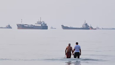 Two men walking hand in hand in the sea with ships in the background.