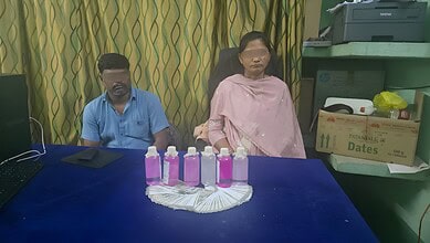 Two individuals seated at a table with pink bottles and a fan in front of them.