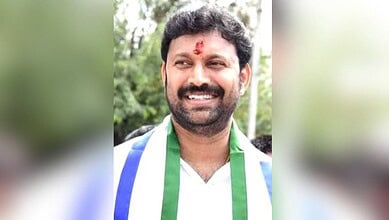 Smiling man with a red tilak on forehead, wearing a white shirt and green scarf.