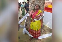 Woman in yellow and red traditional attire leaning against a tree during a cultural event.