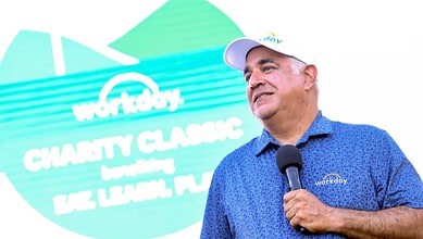 Workday CEO speaking at charity event with a microphone, wearing a blue polo shirt and cap.