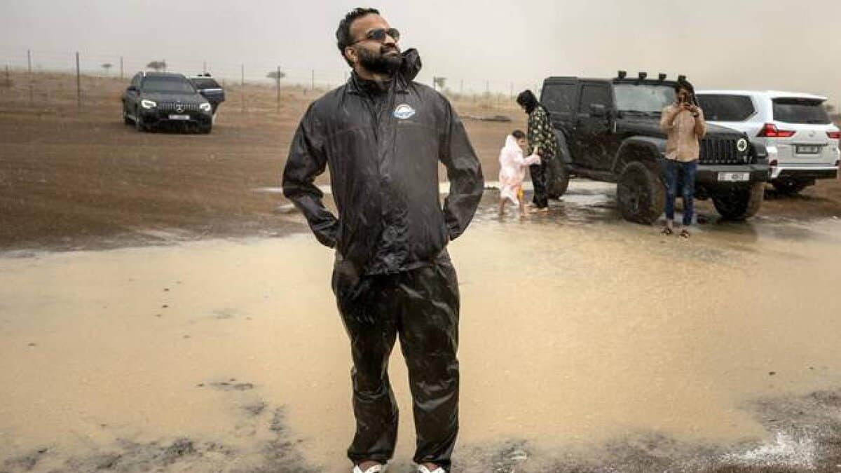 Muhammed Sajjad, known locally as “The Weatherman”, stands in a puddle wearing a black raincoat and flip-flops during rain in the Sharjah desert, with off-road vehicles and other participants in the background.