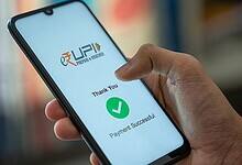 A person holding a smartphone displaying a successful UPI payment confirmation screen. The screen shows the UPI logo, a green checkmark, and the text "Thank You" and "Payment Successful" in English.