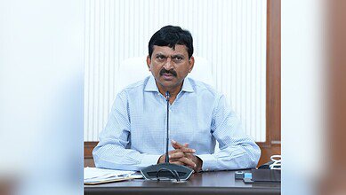 A man with a mustache wearing a light blue checkered shirt sits at a desk speaking into a microphone. He is gesturing with his hands while speaking. A conference phone and papers are visible on the desk in front of him, with a white curtained window in the background.