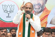 Bandi Sanjay in traditional white and saffron attire speaks passionately at a BJP event, gesturing with his finger raised while standing before the party's lotus symbol.