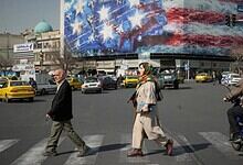 People crossing a busy street in Iran with a large American flag and protest banners in the background.