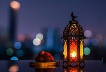 Lantern with intricate design and glowing light, placed beside dates on a balcony at dusk.
