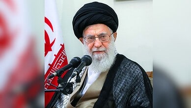 Elderly man in traditional attire speaking at a press conference with Iranian flag in background.