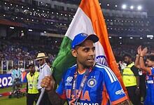 Suryakumar Yadav holding the Indian flag after a cricket match victory, surrounded by teammates and fans.