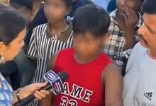 Young boy in red shirt speaking passionately to media surrounded by adults and children.