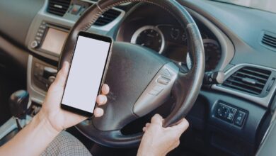 Image shows A driver holding a smartphone with a blank screen while steering a car with their other hand.