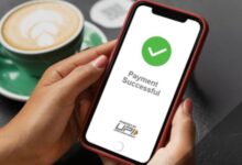 Image shows A hand holds a smartphone displaying a "Payment Successful" confirmation screen powered by UPI, with a coffee cup in the background.