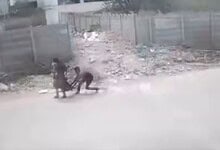 People walking along a dusty, unpaved road with scattered debris and fencing in the background.