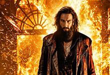 A man with long hair and a beard standing in front of a fiery background with sparks and flames.