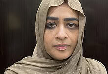 A woman wearing a beige hijab with a serious expression, indoors against a dark background.