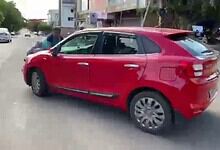 A red hatchback car parked on a city street with pedestrians and buildings in the background.