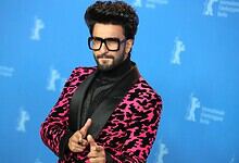 A man with dark hair and beard wearing black glasses and a pink and black patterned blazer at a media eve.