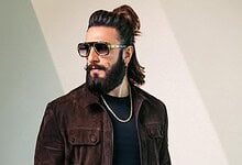 Man with long hair, beard, and sunglasses wearing a dark jacket and gold chain.