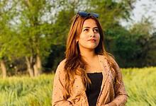 Sunlit woman with long brown hair standing in a lush green park during daytime.