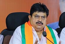 A man speaking at a press conference, wearing a white shirt and a tricolour sash, with a serious expressi.