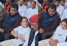 A young girl in white traditional attire sitting next to a man in a red cap and white kurta, with several.