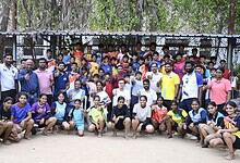 Group of young men and women posing outdoors in front of a chain-link fence, smiling and making gestures.